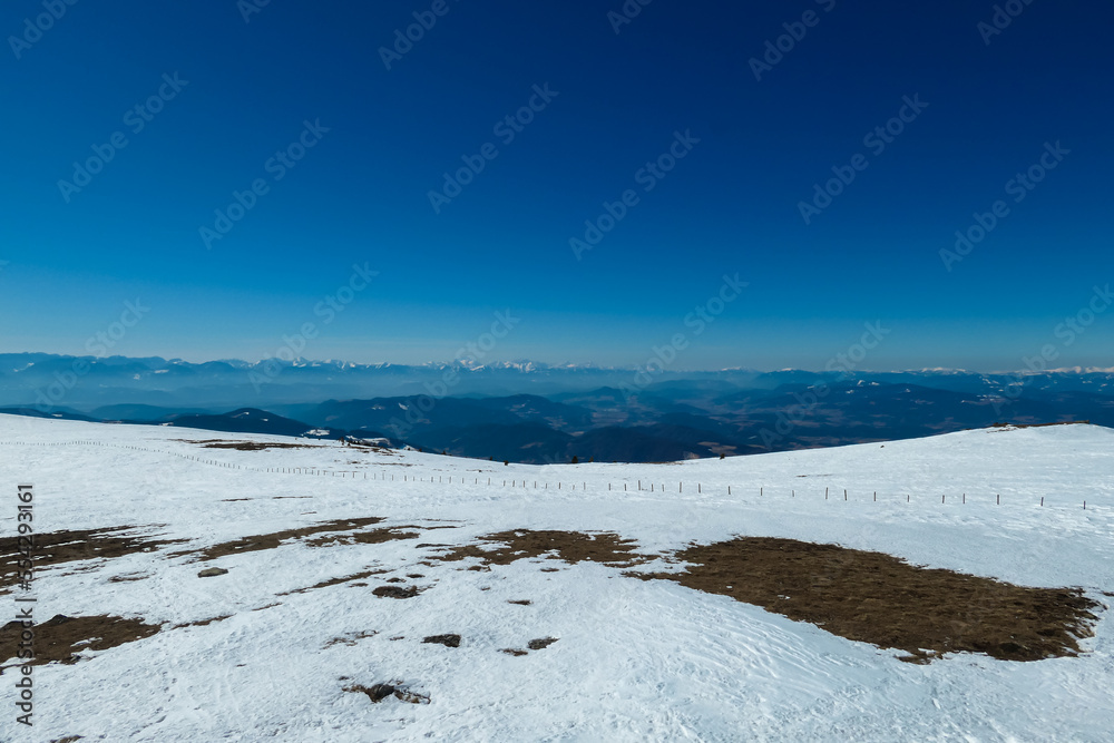 Panoramic view on summit cross of mountain peak Grosser Sauofen in winter on Saualpe, Lavanttal Alps, Carinthia, Austria, Europe. Snowcapped mountain ranges of Karawanks and Kamnik Savinja Alps