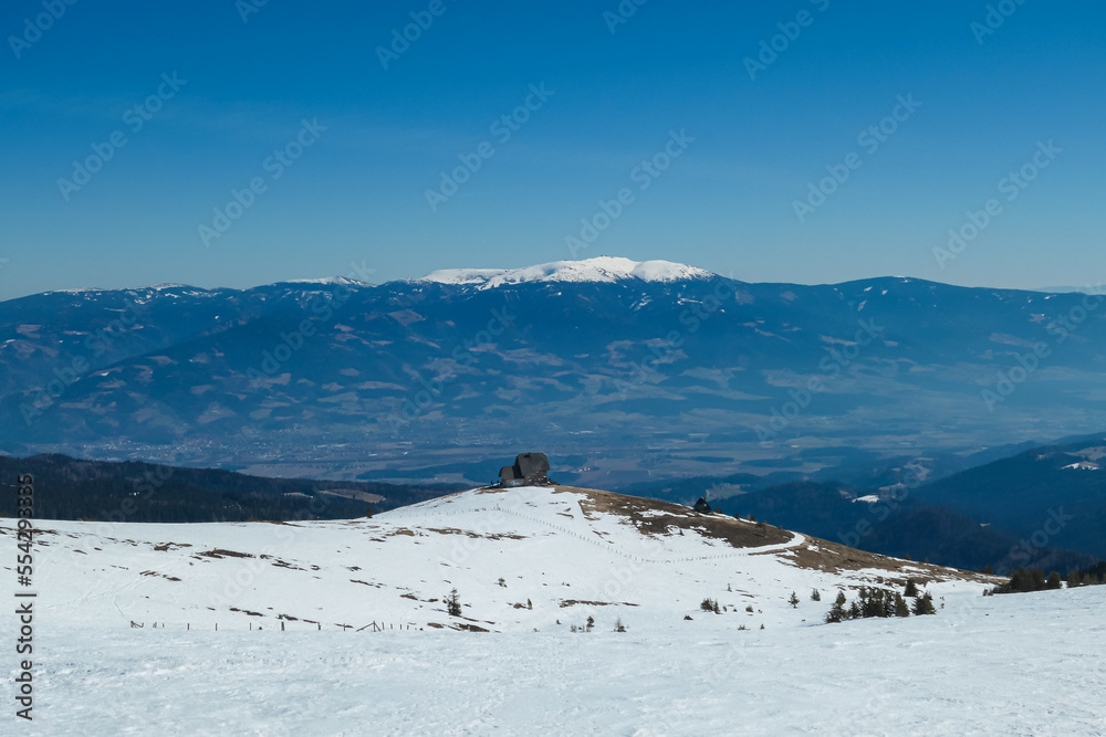 Panoramic view of mountain hut Wolfsbergerhuette (Wolfsberger Huette) on Saualpe, Lavanttal Alps, Carinthia, Austria, Europe. Looking at snow covered Koralpe. Alpine road leading to remote cottage