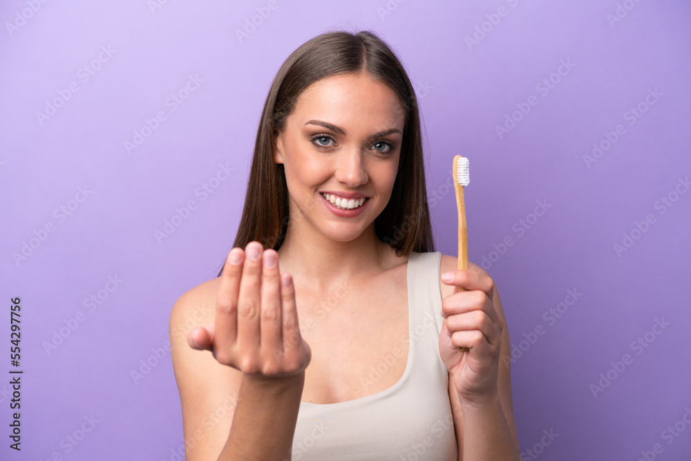 Young caucasian woman brushing teeth isolated on purple background inviting to come with hand. Happy that you came Young caucasian woman brushing teeth isolated on purple background inviting to come with hand. Happy that you came