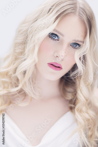 Young woman with blonde curly hair, blue eyes, pink lips and clear skin against a white background. Beauty photoshoot