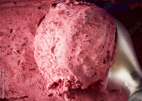 A scoop of cherry ice cream with a textured surface, placed against a backdrop of more frozen dessert.
