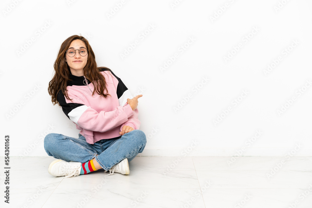 Young caucasian woman sitting on the floor isolated on white background pointing back