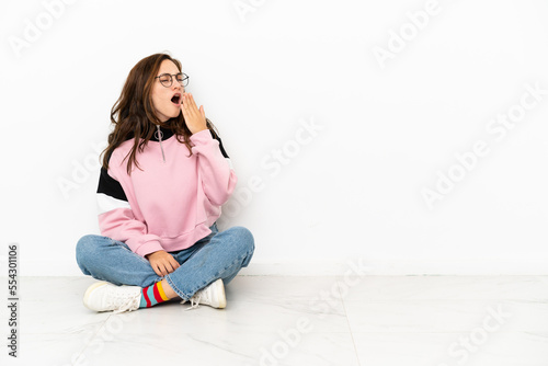 Young caucasian woman sitting on the floor isolated on white background yawning and covering wide open mouth with hand