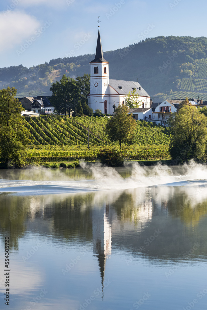 White church with tall steeple reflecting on a river with vineyards along sloped banks and blue sky; Kesten, Germany White church with tall steeple reflecting on a river with vineyards along sloped banks and blue sky; Kesten, Germany