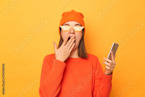 Image of surprised excited woman wearing orange sweater, beanie hat and glasses in minecraft style, holding cell phone in hand, covering mouth with palm, standing isolated over yellow background. photo