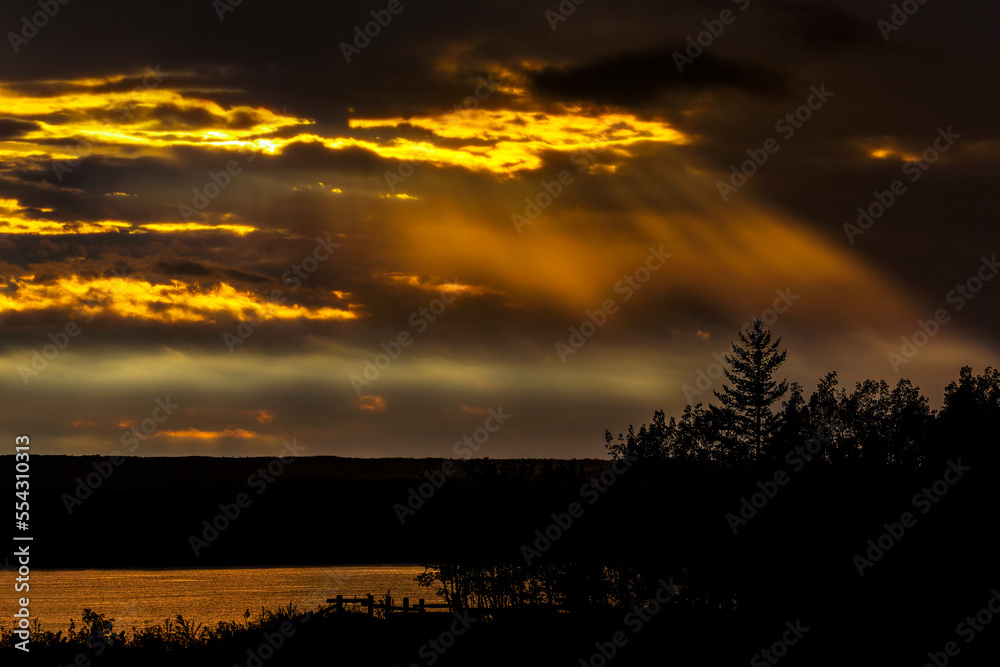 Dramatic colourful sky with sun beams coming down from the sky reflecting on a lake with trees silhouetted in the foreground; Calgary, Alberta, Canada Dramatic colourful sky with sun beams coming down from the sky reflecting on a lake with trees silhouetted in the foreground; Calgary, Alberta, Canada