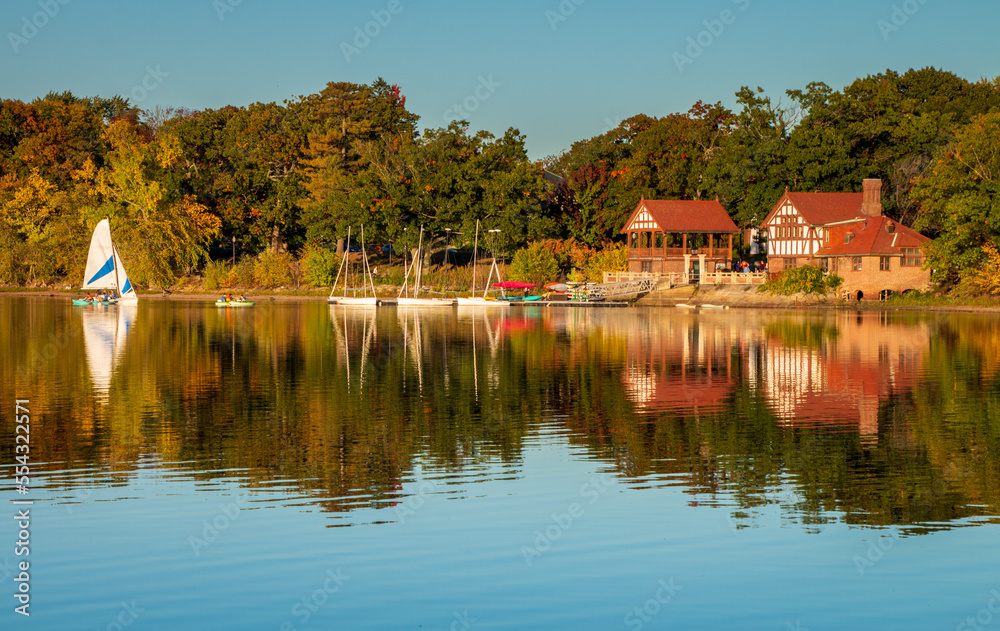 Obraz premium Jamaica Pond view across the water of the boat house and music building. Boats are in the water. A reflection of the buildings are on the water. There is a blue sky. Taken during the golden hour.