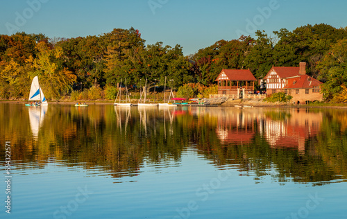 Jamaica Pond view across the water of the boat house and music building. Boats are in the water. A reflection of the buildings are on the water. There is a blue sky. Taken during the golden hour. photo