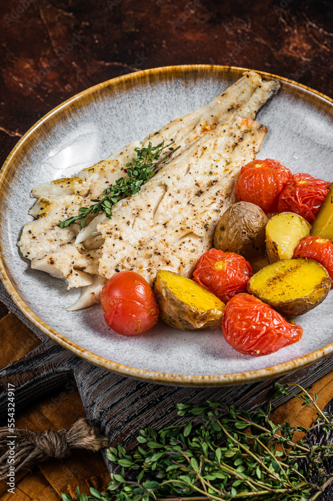 Roasted Pollock fish fillet in plate with garnish tomato and potato. Dark background. Top view