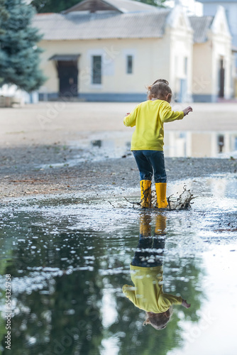 a little girl in yellow clothes and rubber boots cheerfully runs through the puddles after the rain in the warm season on a walk