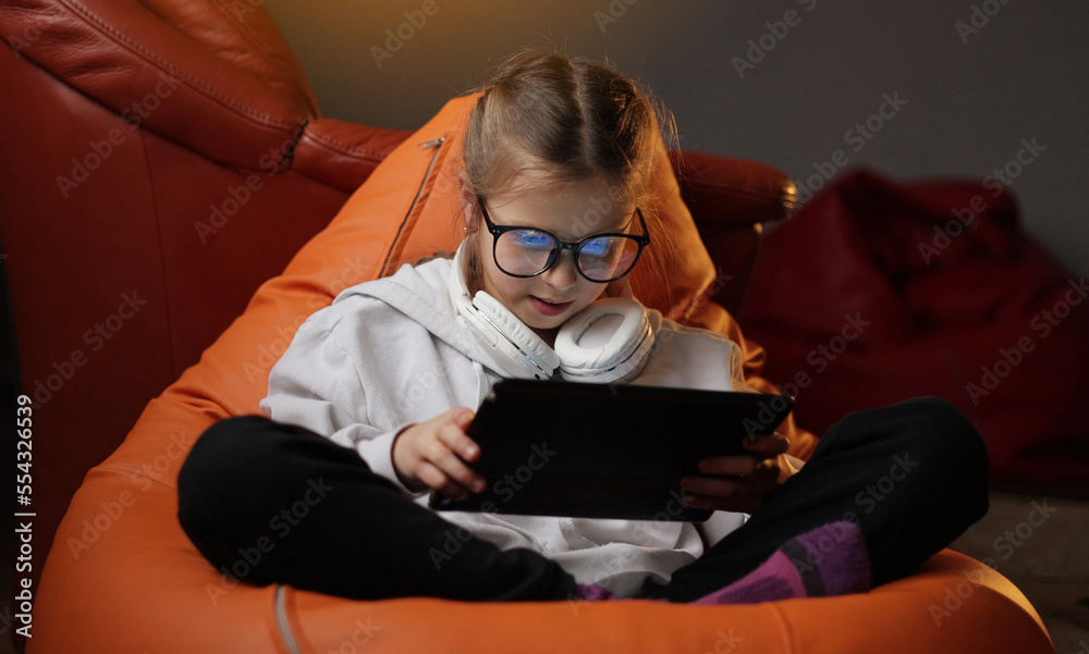 Excited teen girl plays game at home on digital tablet technology device sitting on sofa. Emotional child in headphones and glasses holds pad computer surfing internet. Children tech addiction concept