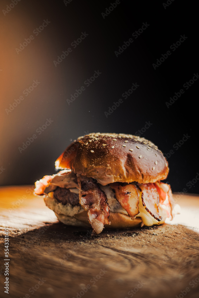 
Tasty beef, cheese, burger with many slices of bacon, warm coming out of smoke on a wooden cutting board on a black background, 
with fire on the stage.  - obrazy, fototapety, plakaty 