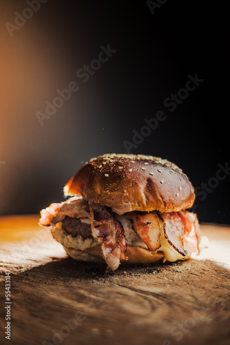 Tasty beef, cheese, burger with many slices of bacon, warm coming out of smoke on a wooden cutting board on a black background, with fire on the stage. 