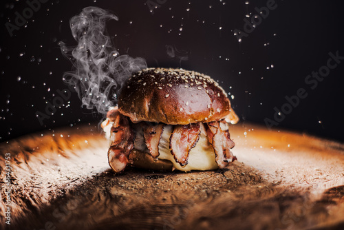  Tasty beef, cheese, burger with many slices of bacon, warm coming out of smoke on a wooden cutting board on a black background.