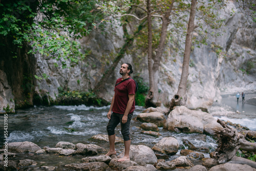A young male tourist near a clear mountain stream in the rocks overgrown with greenery, on a summer day
