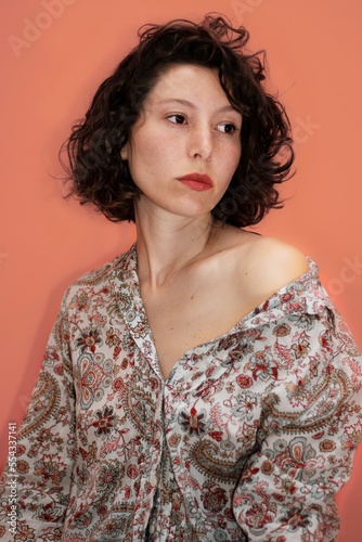 studio shot of a beautiful young woman with curlers and short brown hair posing confidently. The background is pink-orange
