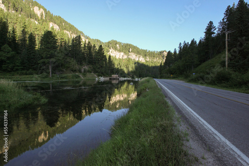 A stream runs parallel open road at the base of a mountain valley.; Deadwood, South Dakota