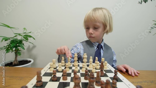 Caucasian boy is playing chess with himself. Focused 5 years old boy in smart suit is larning how to play chess, looking at figures and the board.  photo