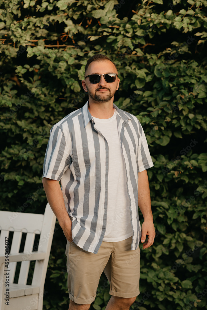 A man is posing in the garden. A guy with green leaves in the background.