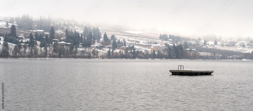 A lonely swimming platform in the middle of a lake in winter, surrounded by fog and snow capped mountains.