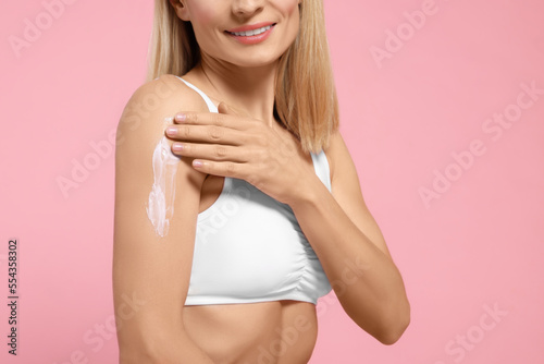 Woman applying body cream onto her arm against pink background  closeup. Space for text