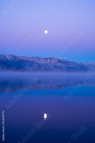 妖しいグラデーションの空の月と山々を湖面に映す朝靄の漂う夜明けの湖。