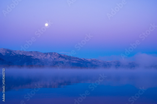 妖しいグラデーションの空の月と山々を湖面に映す朝靄の漂う夜明けの湖。