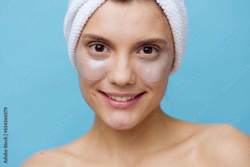 horizontal photo of a joyful, friendly woman standing on a blue background with a towel on her head and patches on her eyes, smiling broadly at the camera horizontal photo of a joyful, friendly woman standing on a blue background with a towel on her head and patches on her eyes, smiling broadly at the camera