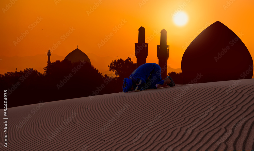 An Arabian man praying on the sand dune with amazing mosque in the background at sunset