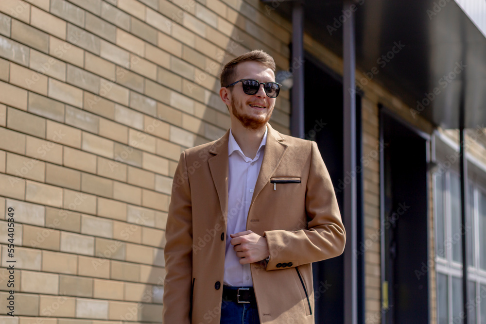 A young man dressed in a beige coat is walking down the street. A young bearded guy with a modern hairstyle and sunglasses on an urban background A young man dressed in a beige coat is walking down the street. A young bearded guy with a modern hairstyle and sunglasses on an urban background