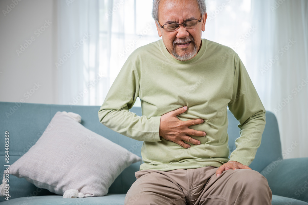 Elderly Asian old man sitting on sofa having stomachache, old age suffering from stomach ache holding his stomach in living room, people health problem, food poisoning