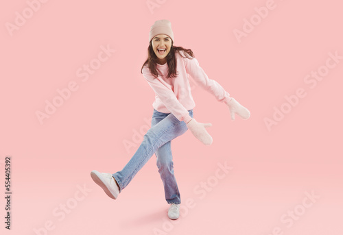 Full body happy lively attractive young woman wearing sweatshirt, jeans, wool winter hat, mittens having fun in pink fashion studio, dancing to music, laughing, doing funny moves with hands and legs