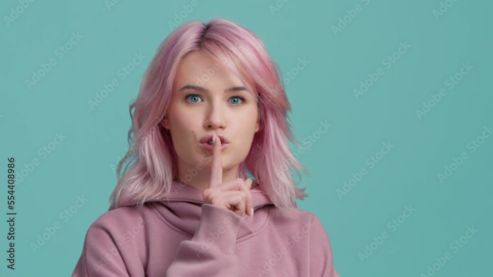 Silence gesture young Hipster girl with pink hair showing index finger to lips. Serious model with trendy hair color makes gesture for silence, looking at camera on blue background. Don't tell gesture Silence gesture young Hipster girl with pink hair showing index finger to lips. Serious model with trendy hair color makes gesture for silence, looking at camera on blue background. Don't tell gesture