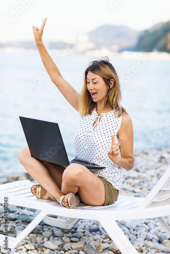 A woman rejoices for making a deal while working at a laptop, sitting on a sun lounger on the seashore, doing online business, combining rest and work