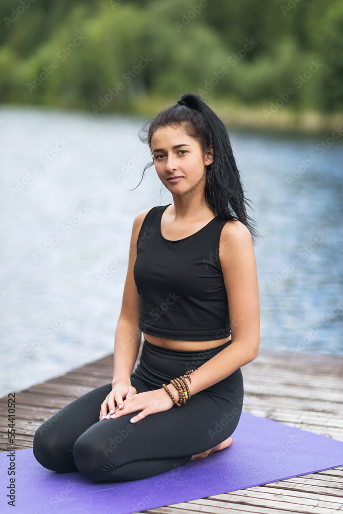 Portrait of a young beautiful female yoga trainer sitting on a wooden footbridge on the shore of a lake