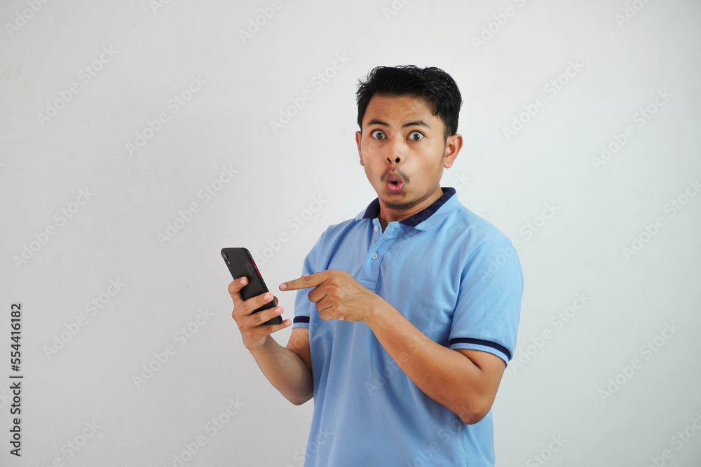 portrait asian man shocked holding phone and pointing at the phone with a finger wearing blue polo t shirt isolated on white background portrait asian man shocked holding phone and pointing at the phone with a finger wearing blue polo t shirt isolated on white background