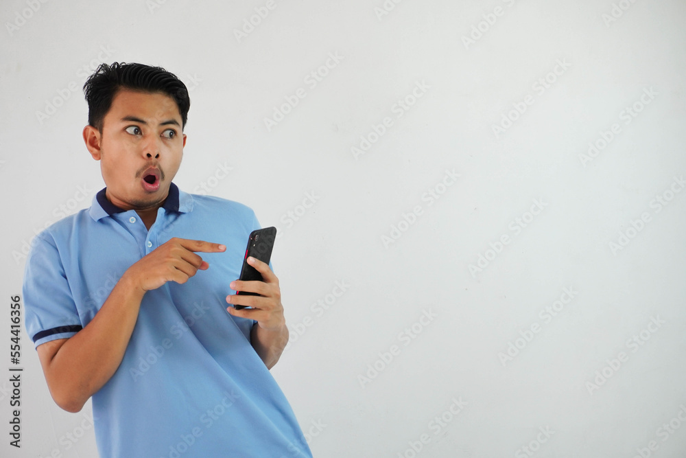 portrait asian man shocked holding phone and pointing at the phone with a finger wearing blue polo t shirt isolated on white background portrait asian man shocked holding phone and pointing at the phone with a finger wearing blue polo t shirt isolated on white background