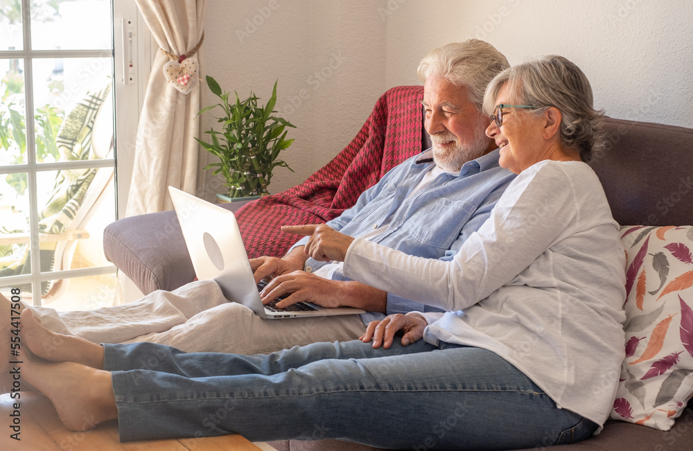Handsome senior couple sitting on sofa at home while browsing on laptop planning a travel destination - smiling caucasian people enjoying retirement