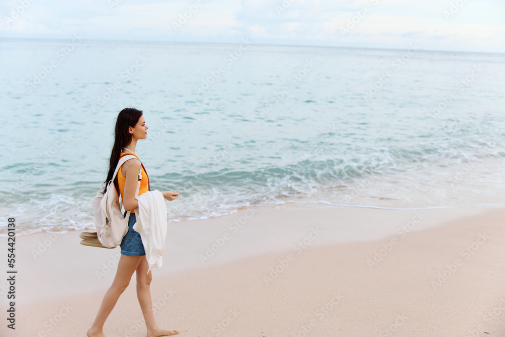 Woman smile with teeth after swimming in the ocean with a backpack in a wet yellow tank top and denim shorts walks along the beach, summer vacation on an island by the ocean in Bali sunset Woman smile with teeth after swimming in the ocean with a backpack in a wet yellow tank top and denim shorts walks along the beach, summer vacation on an island by the ocean in Bali sunset