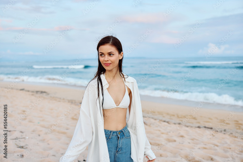 Happy tanned slim woman in white swimsuit shirt and jean shorts walks on the beach on the sand by the ocean with wet hair after swimming, sunset light Happy tanned slim woman in white swimsuit shirt and jean shorts walks on the beach on the sand by the ocean with wet hair after swimming, sunset light