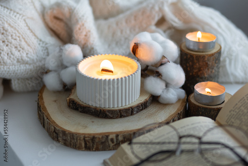 Cozy autumn or winter composition with aromatic candle, wool sweater, cotton, book. Aromatherapy, home atmosphere of cosiness and relax. Wooden background close up.