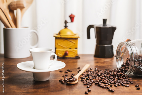 Cup of coffee on a kitchen table with a jug with wooden utensils, a coffee grinder, an Italian coffee maker and an overturned jar with roasted coffee beans, in the background a white curtain.