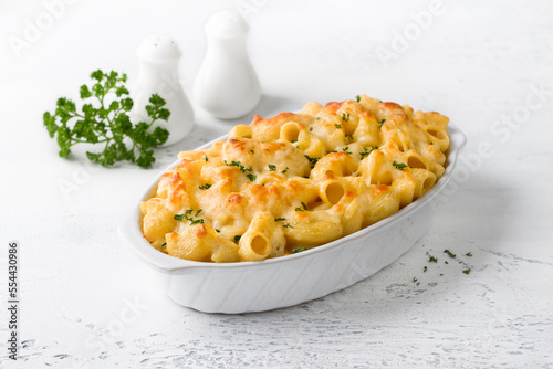 Mac and cheese, baked American pasta in cheese sauce decorated with parsley in a baking dish on a light gray background. Delicious homemade food