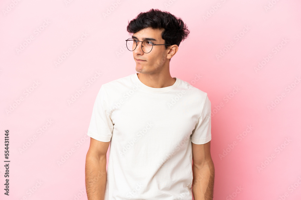 Young Argentinian man isolated on pink background having doubts while looking side Young Argentinian man isolated on pink background having doubts while looking side