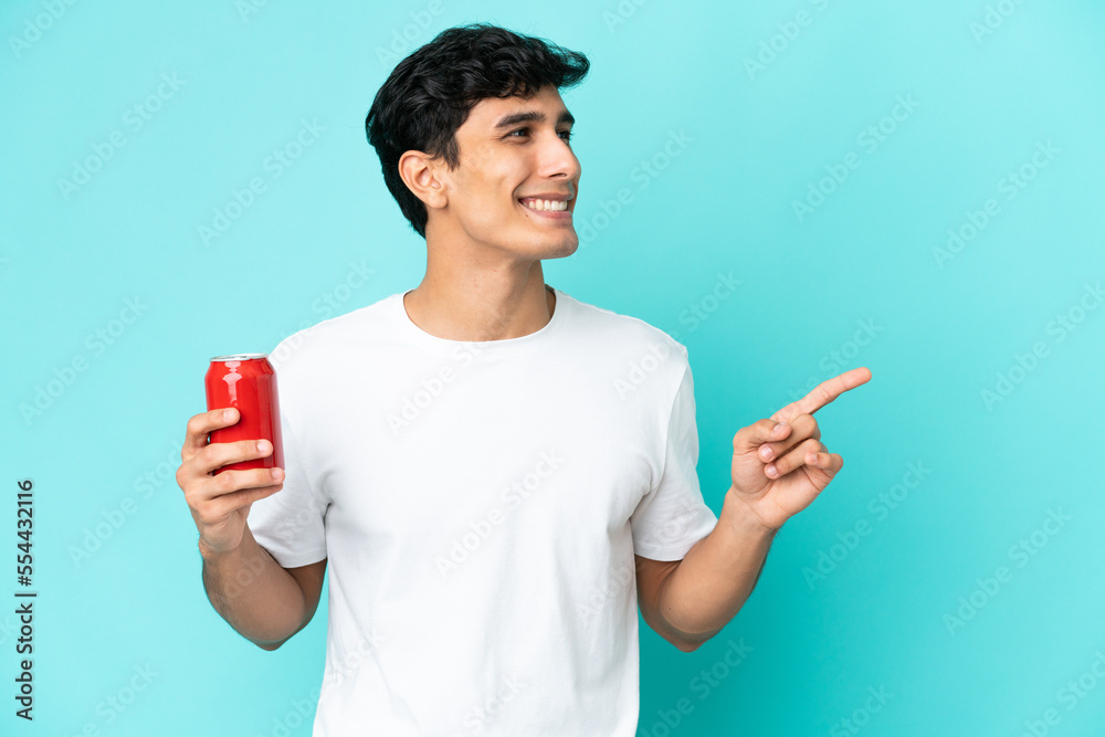 Young Argentinian man holding a refreshment isolated on blue background pointing finger to the side and presenting a product Young Argentinian man holding a refreshment isolated on blue background pointing finger to the side and presenting a product