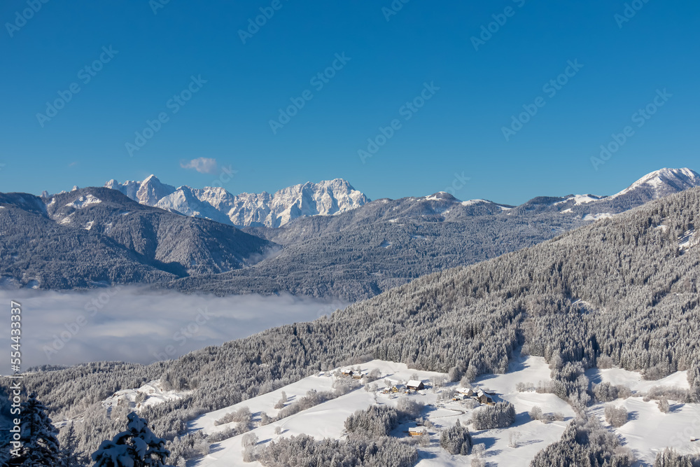 Panoramic view of the snow capped mountain range of Julian Alps seen from Kobesnock near Bad Bleiberg, Carinthia, Austria, Europe. Remote village in winter wonderland on sunny day. Valley full of fog