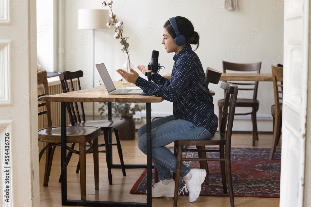 Professional Indian radio presenter woman in headphones speaking on air, sitting at work table with laptop, big mic, broadcasting from home office, empty co-working space. Side full length shot