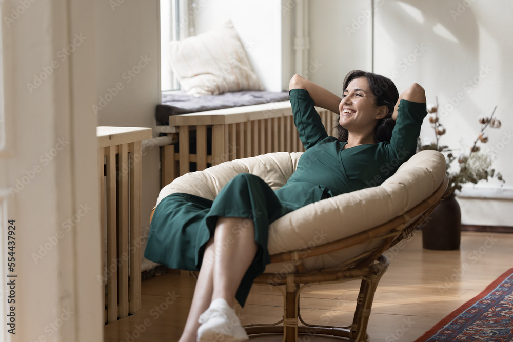 Happy carefree pretty Asian woman relaxing in soft round armchair, resting, holding nap, taking deep breath, looking away, enjoying leisure, comfort at home, thinking, dreaming, smiling, laughing