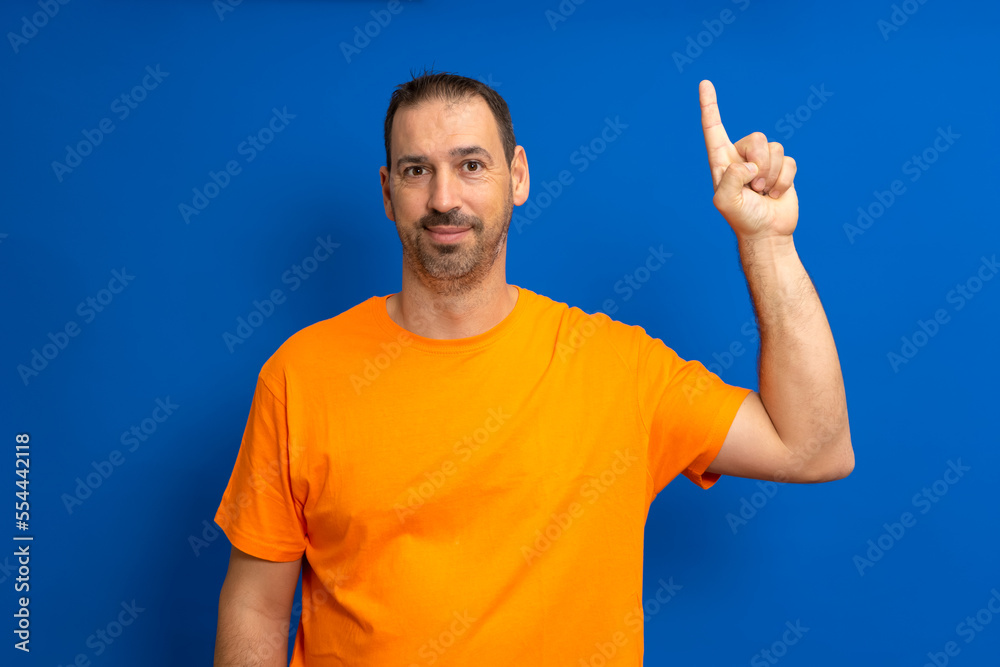 Insightful smart proactive caucasian man 40s in orange t-shirt holding index finger up with great new idea isolated on blue background studio portrait. People lifestyle concept Insightful smart proactive caucasian man 40s in orange t-shirt holding index finger up with great new idea isolated on blue background studio portrait. People lifestyle concept
