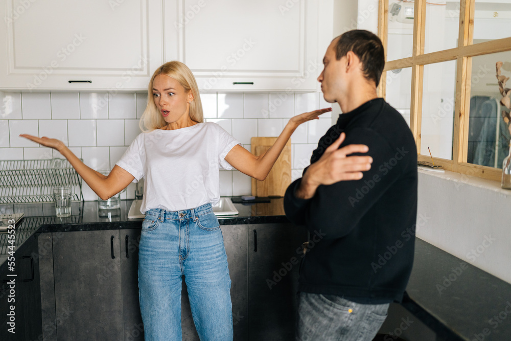 Obraz premium Tired frustrated Caucasian husband ignoring angry blonde wife arguing blaming upset man of problems standing in kitchen. Jealous woman shouting at sad boyfriend. Concept of family problems